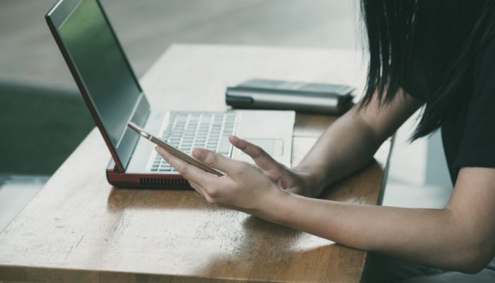 femme-bras-penché-sur-table-tenant-smartphone-avec-ordinateur-ouvert-sur-table